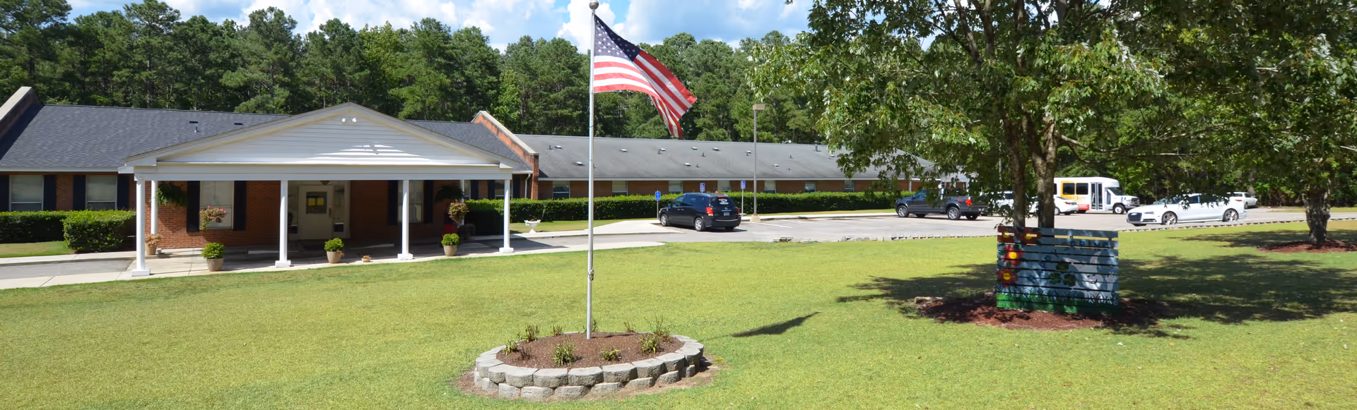 Front exterior of a single-story brick retirement facility with a covered entrance, an American flag on a grassy lawn, parked cars and surrounding trees.