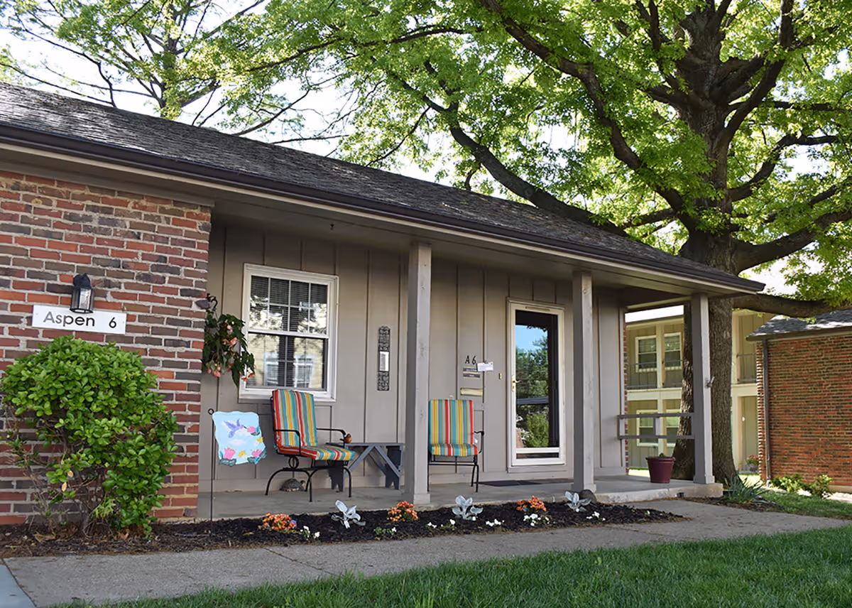 A small covered front porch of a brick-and-siding senior living building with chairs and plants