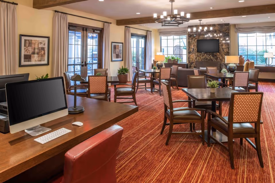 A well-lit common area with multiple tables and chairs arranged on a red patterned carpet. There is a desk with a computer, keyboard, mouse, and lamp in the foreground. The room features large windows with beige curtains, framed artwork on the walls, and a stone fireplace with a mounted flat-screen TV. Several potted plants and table lamps add to the cozy atmosphere.