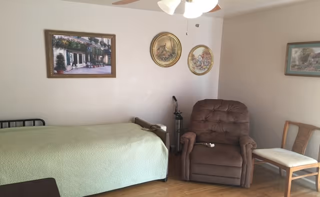 A small bedroom in a senior care home with a bed, recliner, and framed wall art.