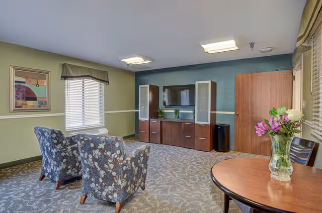 Communal living room with two patterned armchairs facing a wall-mounted TV and media cabinet, plus a small round table with a vase of flowers.