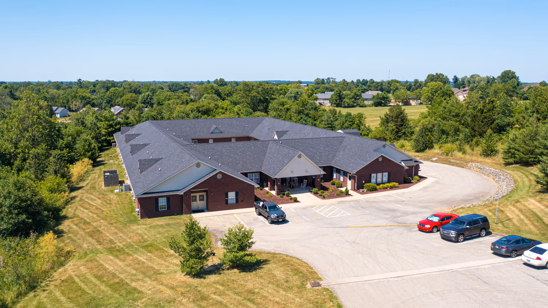 Aerial view of a single-story brick assisted living facility with a dark roof, surrounded by green lawns and trees. Several cars are parked in the parking lot in front of the building. The facility is situated in a suburban area with more houses and greenery in the background under a clear blue sky.