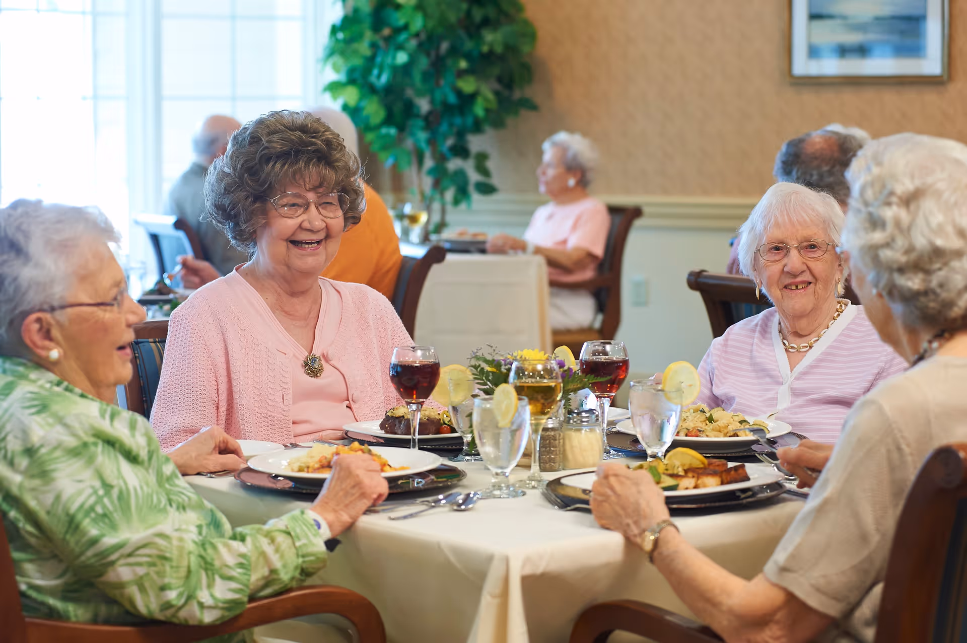 Four elderly women sitting around a dining table in a senior living facility, enjoying a meal together with plates of food and glasses of water and wine. The room has a warm, inviting atmosphere with other residents dining in the background.