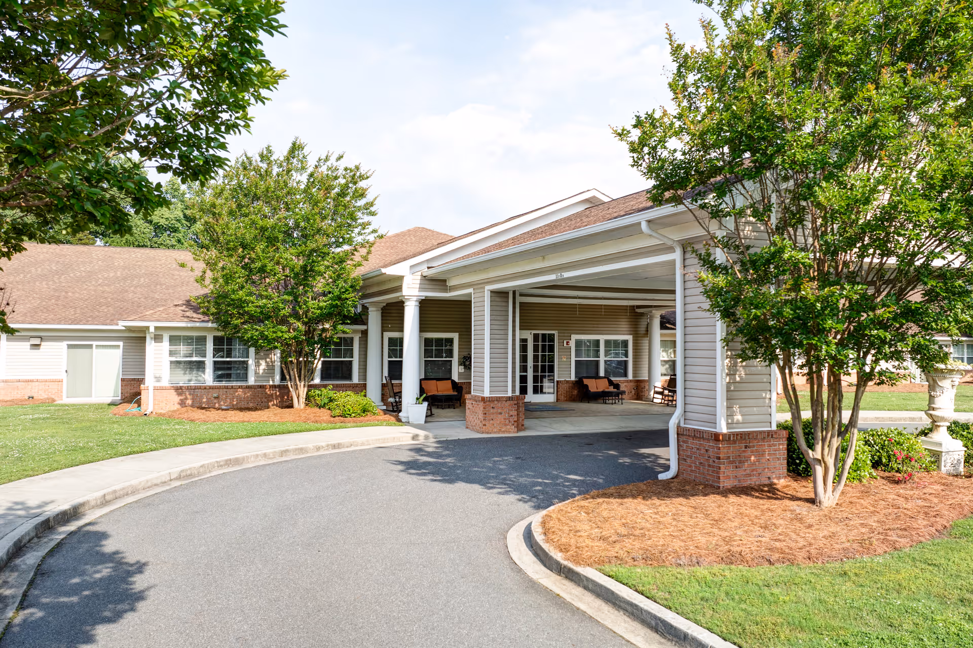 Exterior view of TerraBella Rock Hill facility entrance with a covered drop-off area, surrounded by green trees and well-maintained landscaping under a partly cloudy sky.