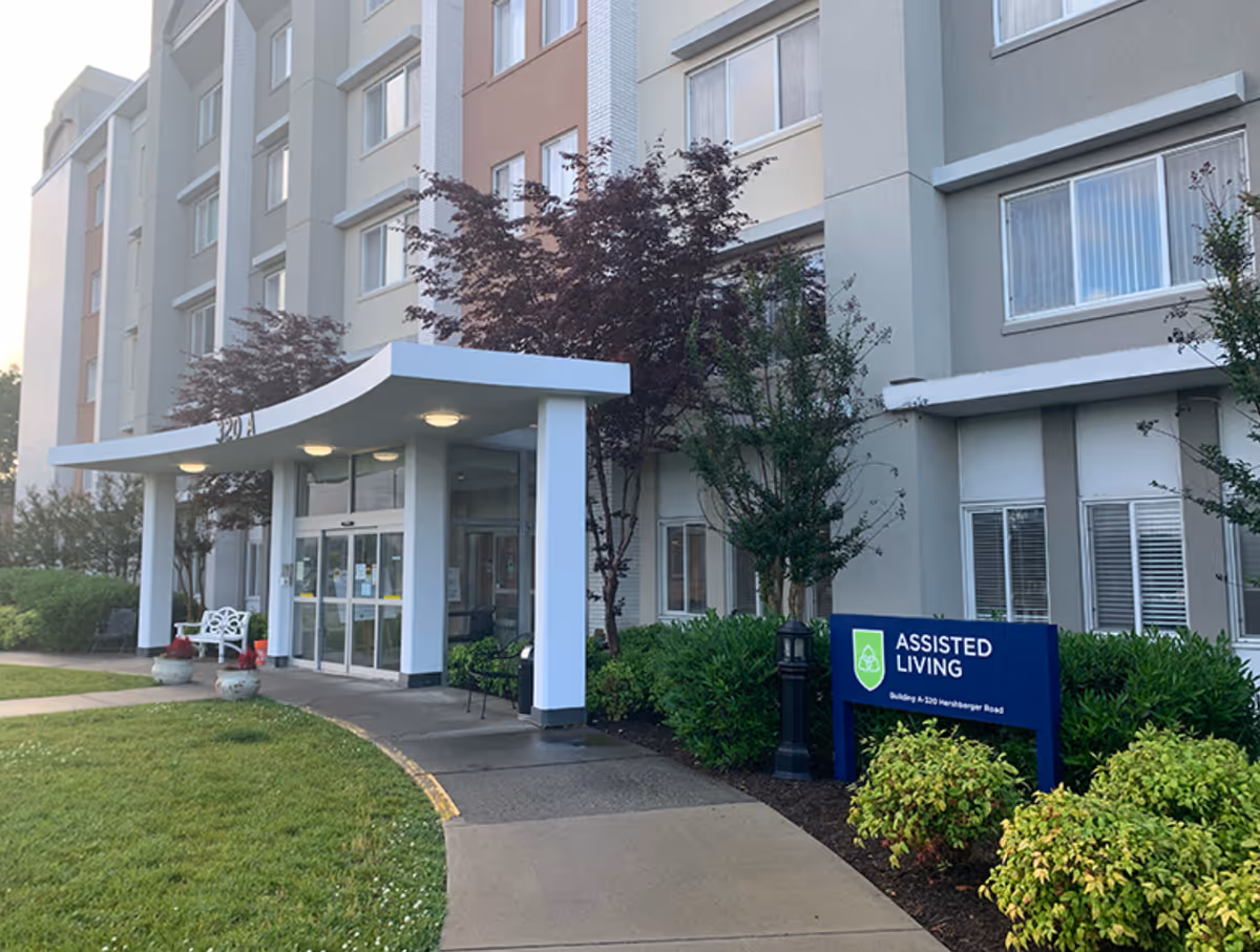 Front entrance of a multi-story assisted living building with a covered canopy, glass doors, landscaping, and an 'Assisted Living' sign.