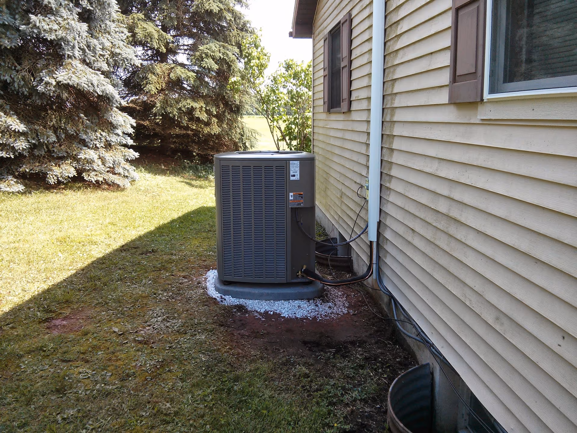 A residential air conditioning unit sits outside a beige siding house with trees nearby.