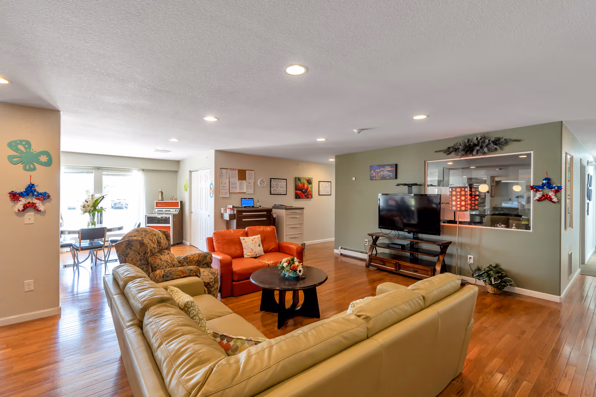 Spacious senior living lounge with beige sectional, orange loveseat, coffee table, and wall-mounted TV.