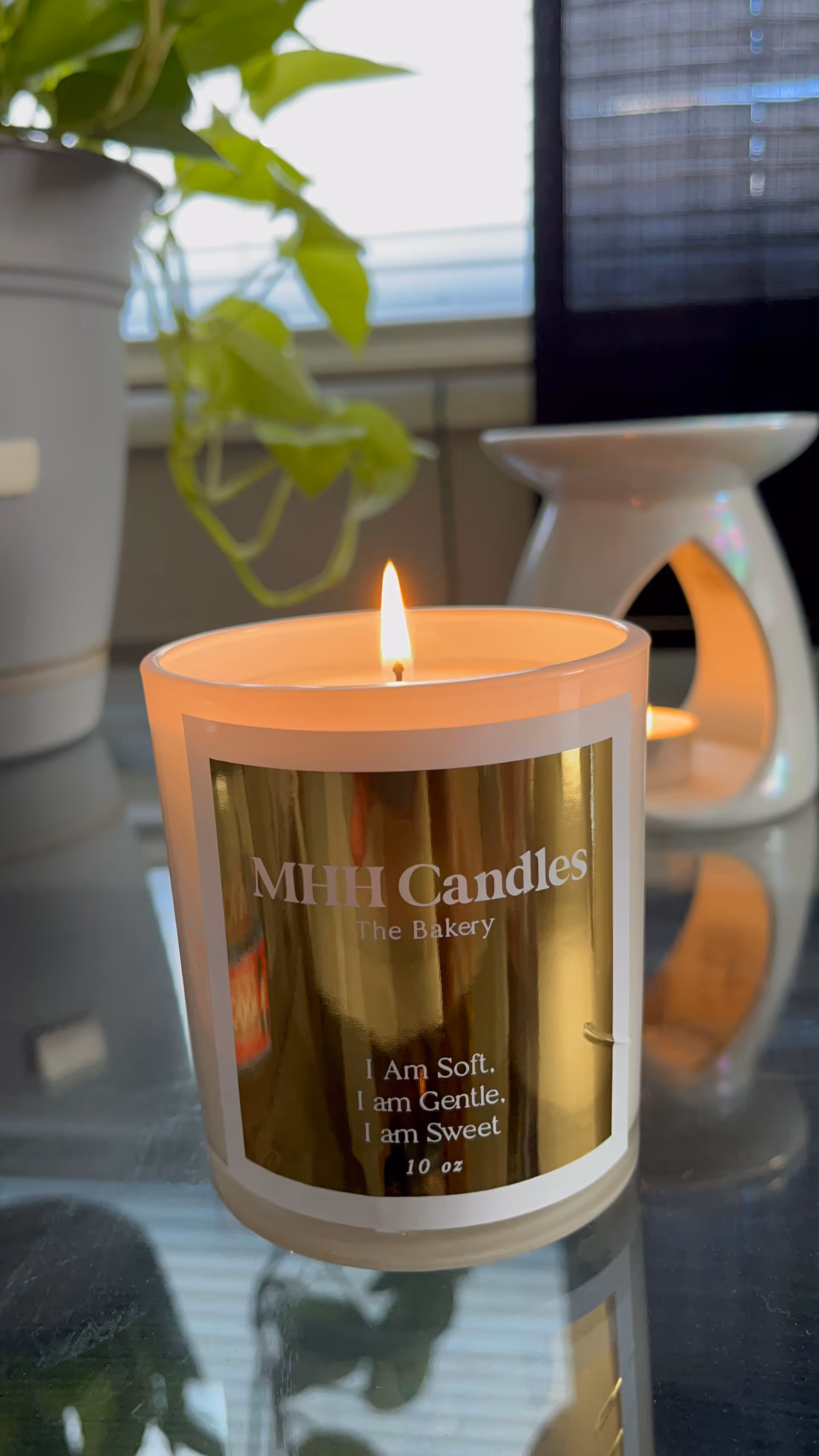 Lit candle labeled MHH Candles sits on a glass table beside a plant.