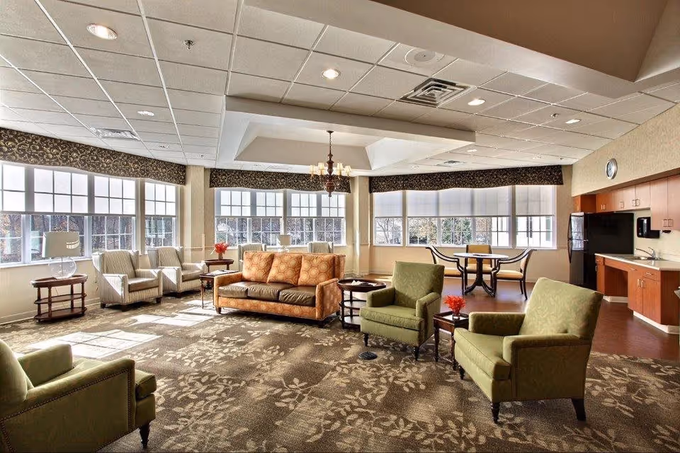 Sunlit communal living room with sofas, armchairs, side tables and a small kitchenette/dining area under large windows.