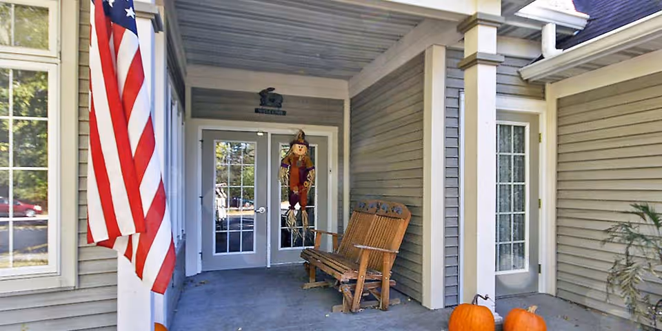 Covered front entrance with double glass doors, an American flag, a wooden bench, and pumpkins on the porch.