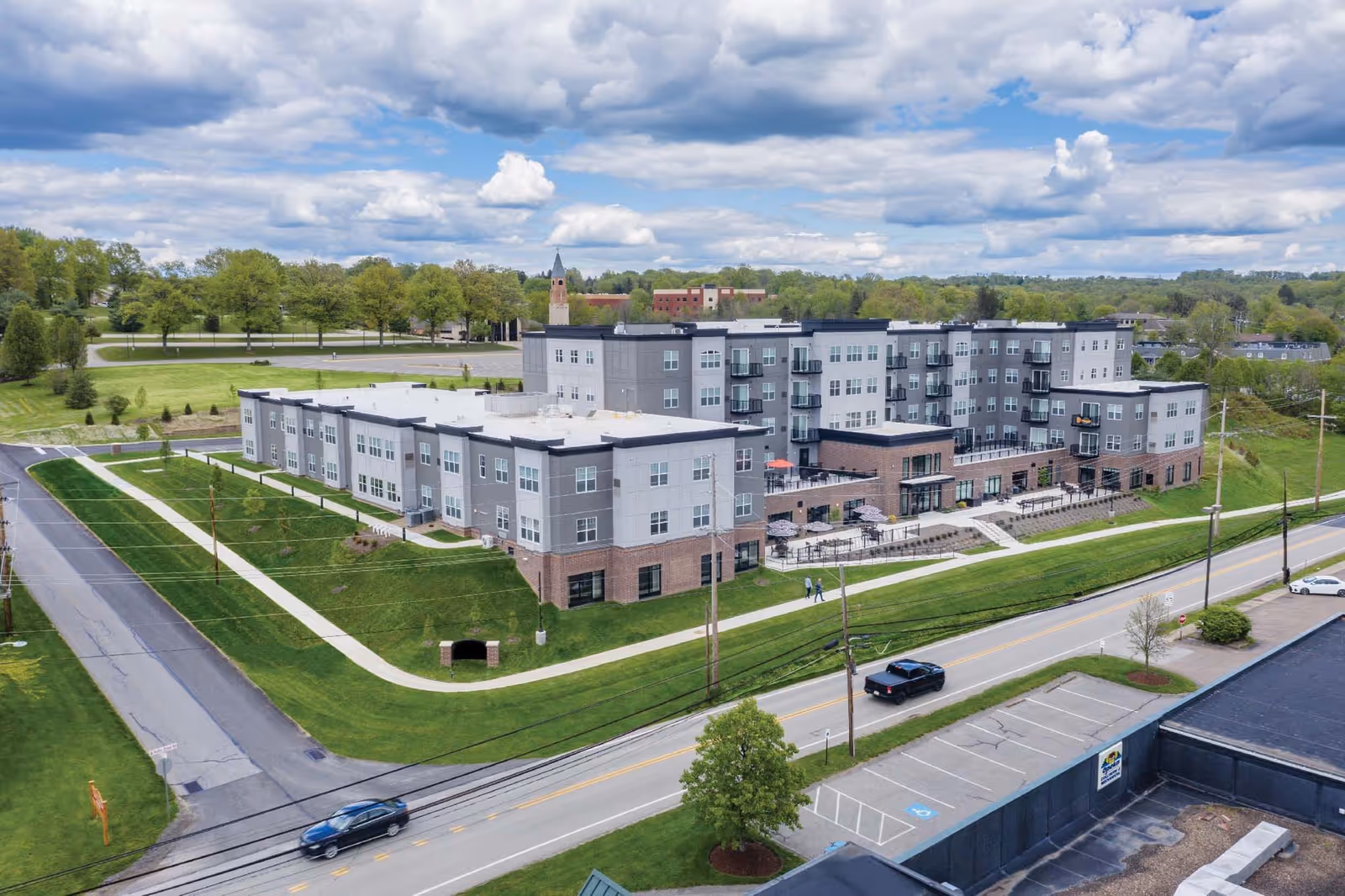 Aerial view of a large, modern senior living facility named Ridgecrest of McMurray. The building is multi-story with gray and brick exterior walls, balconies, and outdoor seating areas. Surrounding the building are well-maintained green lawns, sidewalks, and nearby roads with a few cars. The sky is partly cloudy with patches of blue.