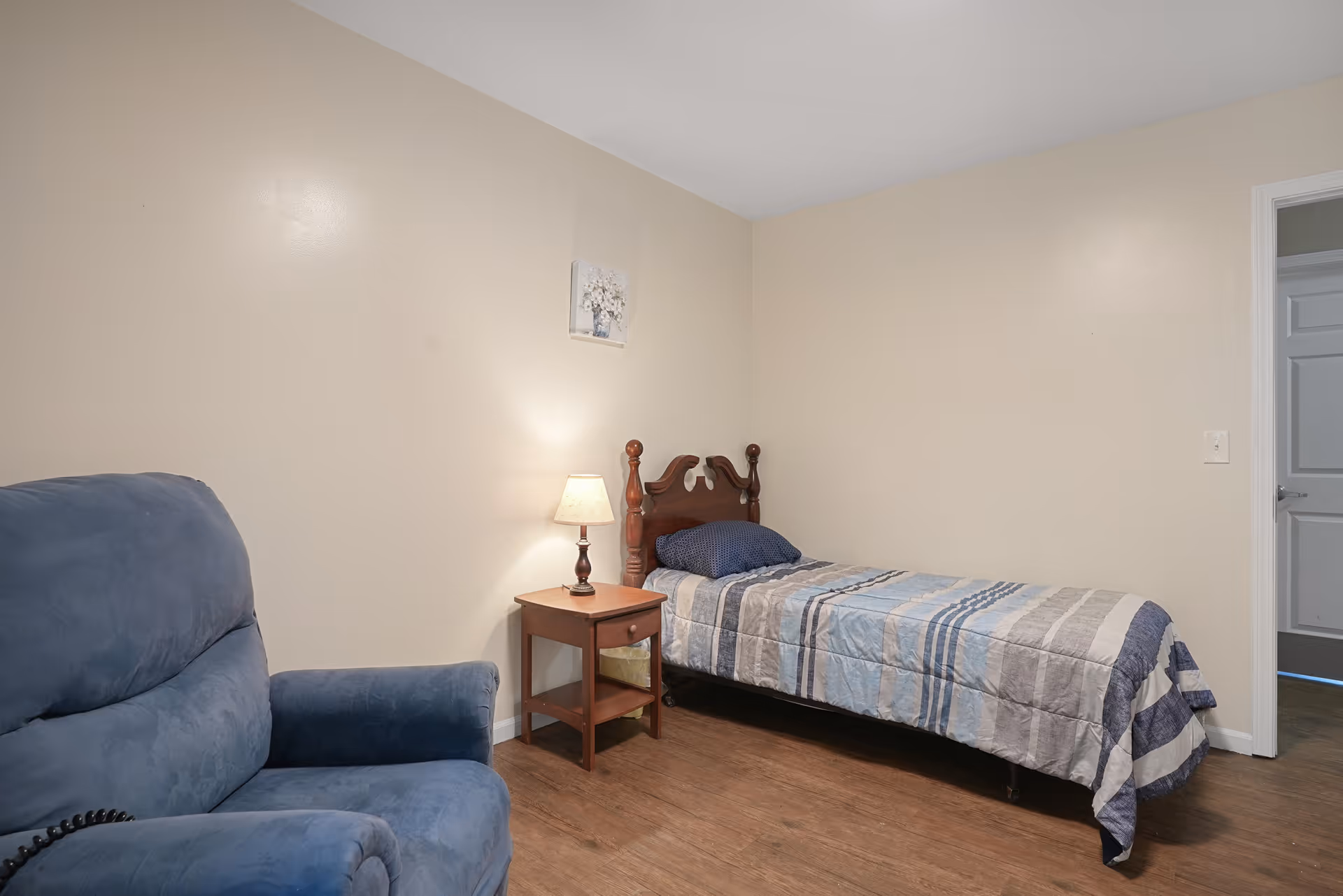 A simple bedroom with a single bed covered in a striped blue and gray comforter, a wooden nightstand with a lamp, a blue upholstered armchair, and beige walls with a small framed picture above the bed.