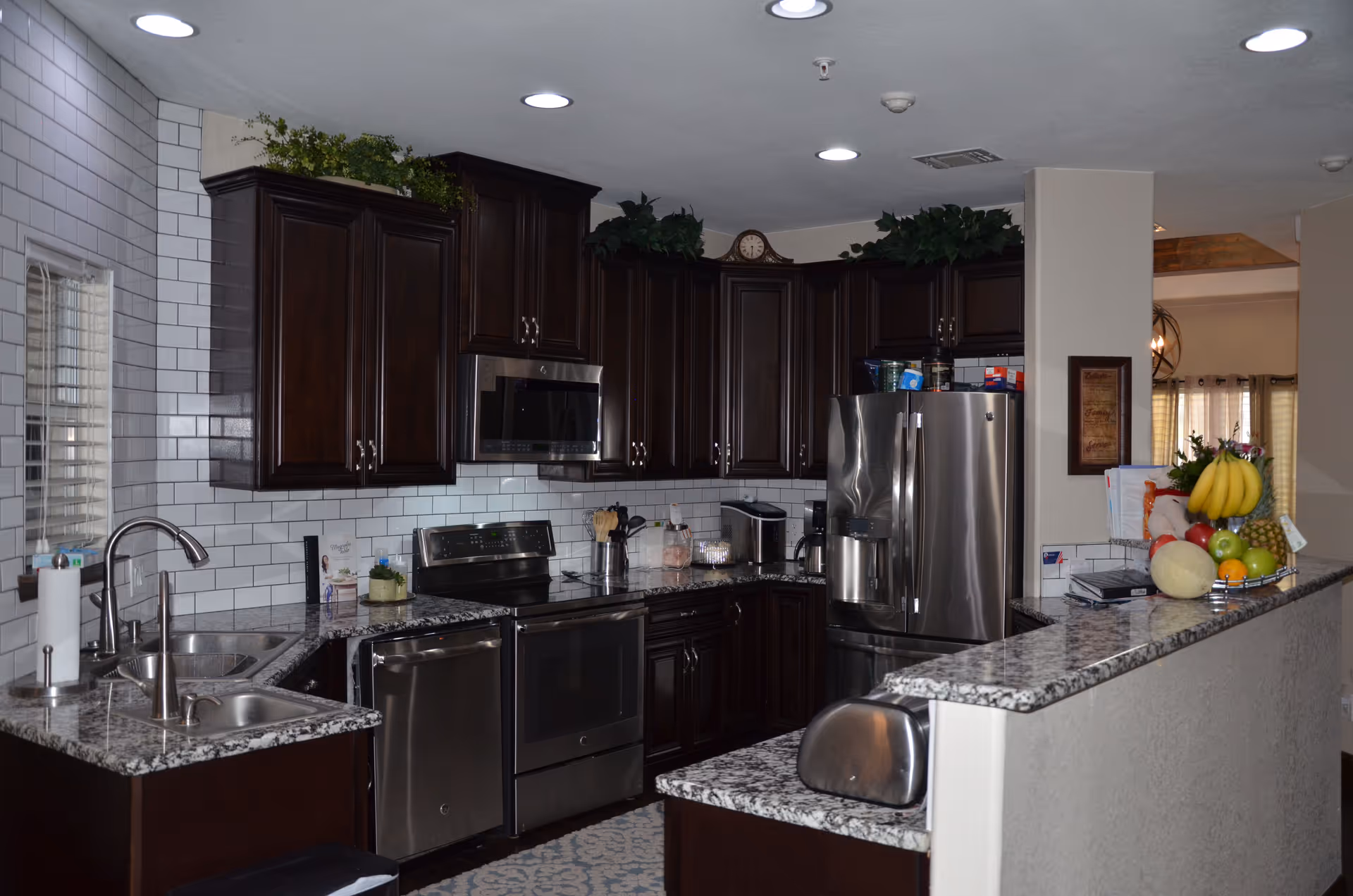 Modern kitchen with dark wooden cabinets, stainless steel appliances including a refrigerator, microwave, oven, and dishwasher. The countertops are granite with a double sink and a paper towel holder. There are decorative plants on top of the cabinets and a fruit basket with bananas, apples, and other fruits on the counter. The backsplash is white subway tile, and the kitchen is well-lit with recessed ceiling lights.