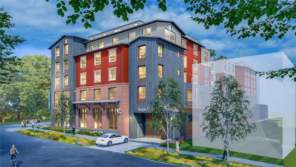 Exterior view of a multi-story senior living facility named Aegis Living Ballard, featuring a modern design with red and gray siding, large windows, and a brick base. The building is surrounded by trees and landscaping, with a few people walking and a car parked near the entrance.