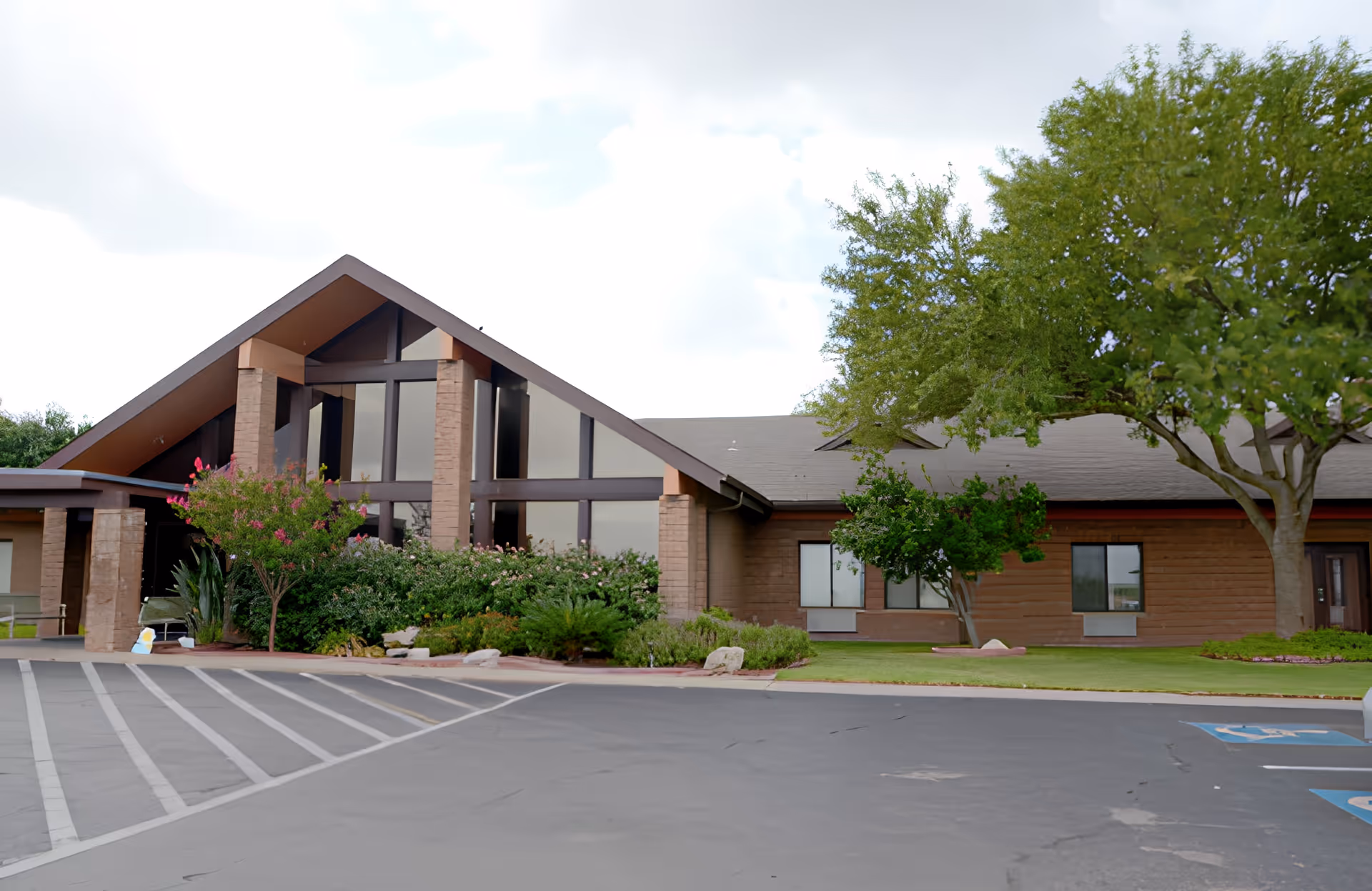 Front exterior of a single-story senior living facility with an A-frame entrance, large windows, landscaped shrubs, and a parking lot.