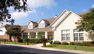 Exterior view of a single-story senior living facility building with a stone facade, large windows, and a covered porch with white columns. The building is surrounded by well-maintained landscaping including bushes, small trees, and potted plants. The sky is clear with a few clouds.