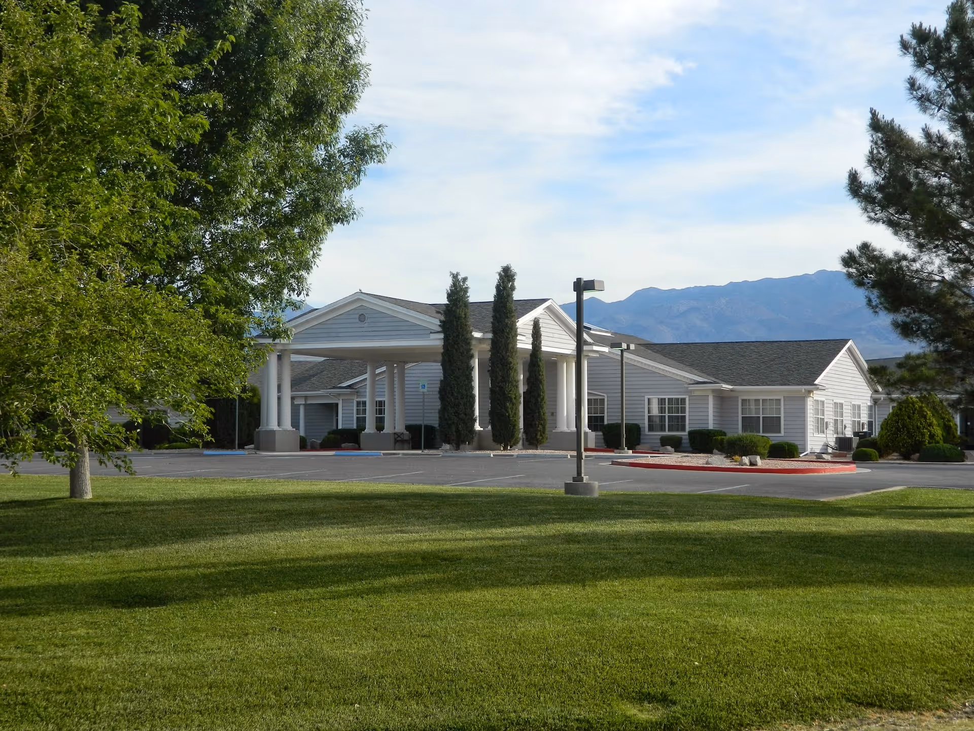 Exterior view of a single-story building with a covered entrance, surrounded by green grass, trees, and shrubs, with mountains visible in the background under a partly cloudy sky.