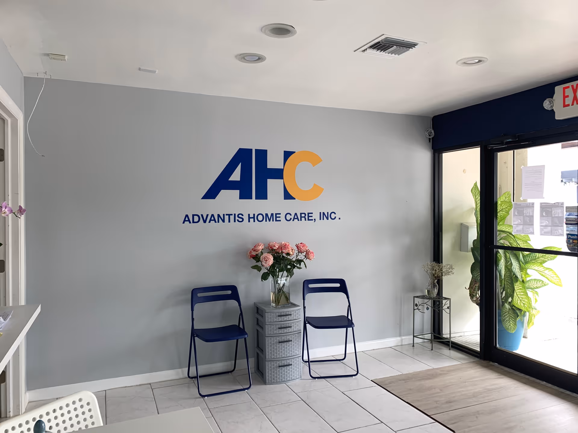 Lobby area with Advantis Home Care logo on wall, blue chairs, glass door, and plants.