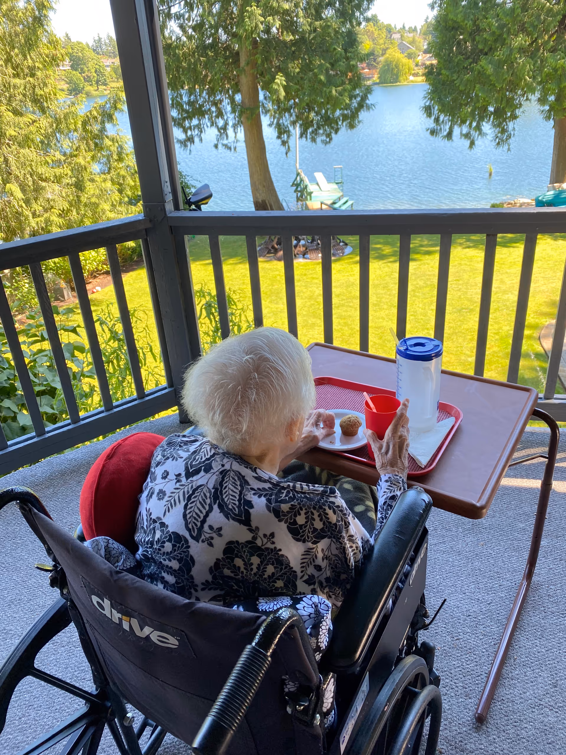 An elderly person in a wheelchair sits at an outdoor balcony table overlooking a lake.