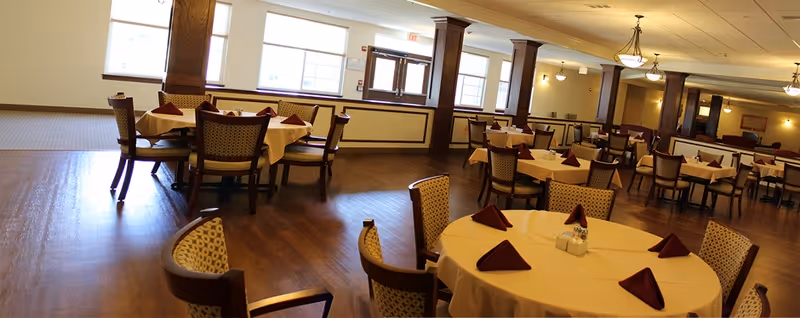 Spacious dining room with round tables set with tablecloths and folded napkins, wooden chairs, and hardwood floors.