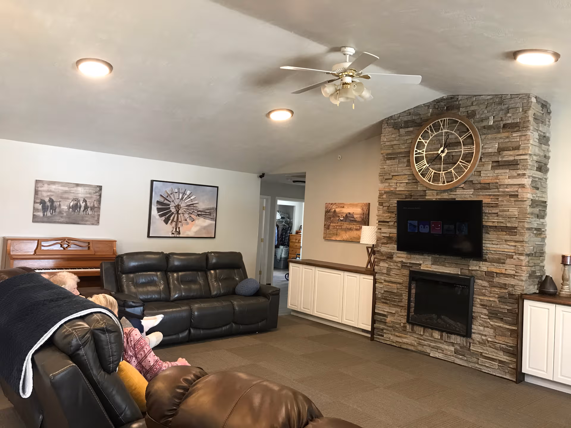 Cozy senior living living room with leather sofas, a stone fireplace, and a ceiling fan.
