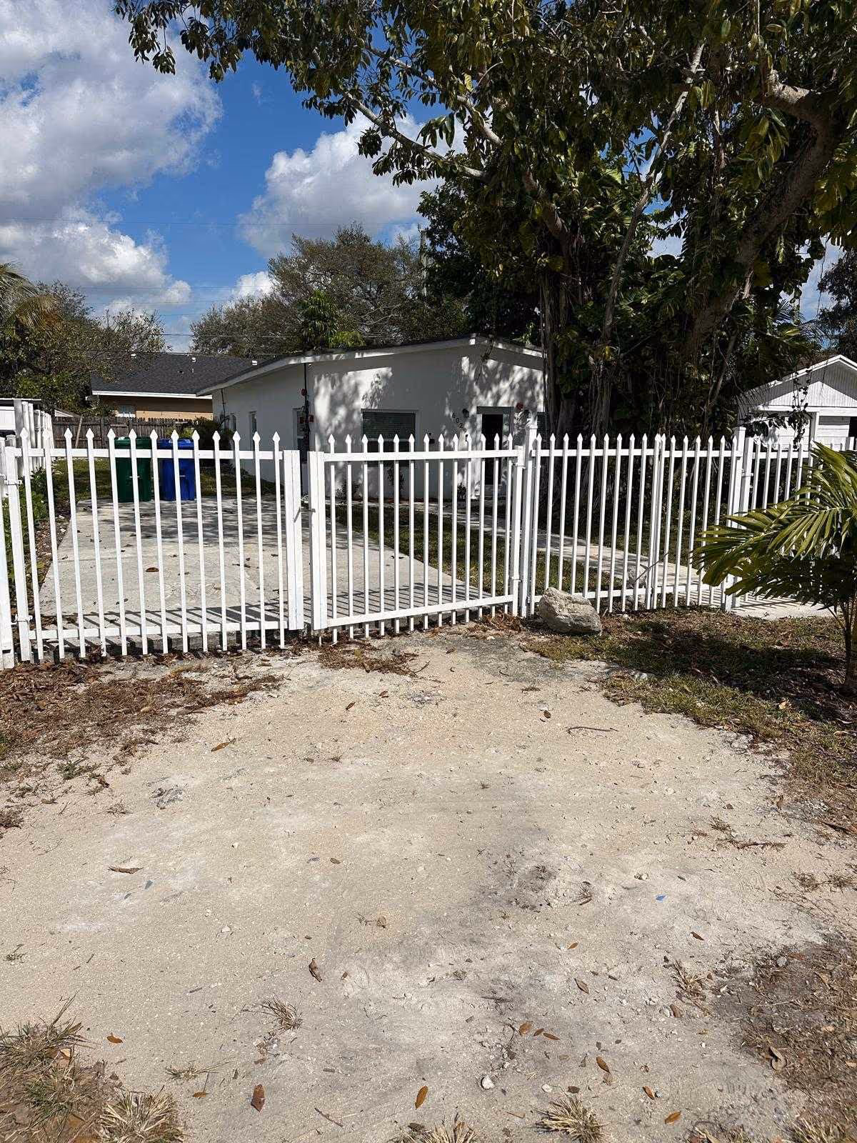 White metal gate opens to a single-story building with trees and a driveway.