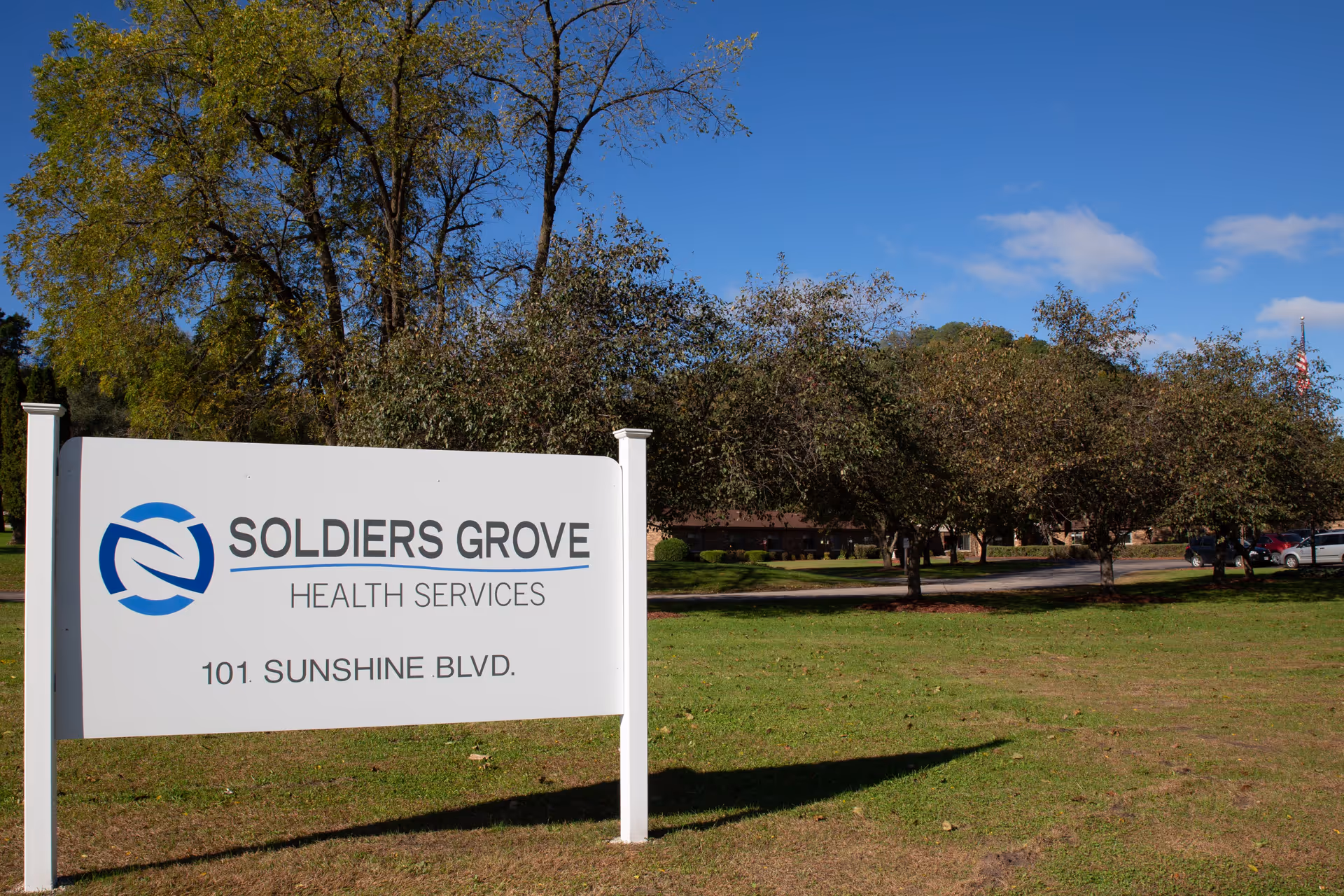 White sign for Soldiers Grove Health Services (101 Sunshine Blvd.) on a grassy lawn with trees and the facility building in the background.
