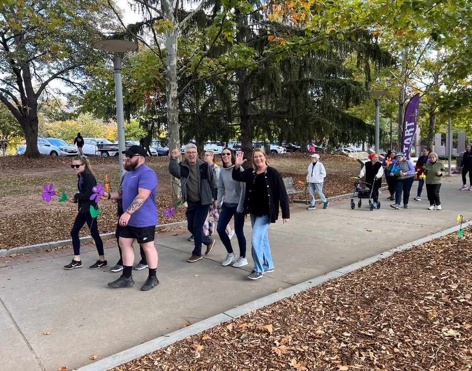 A group of people walking on a paved path in a park-like outdoor setting with trees and parked cars in the background. Some individuals are holding purple flower decorations, and one person is using a walker. The group appears to be participating in a community or social event.