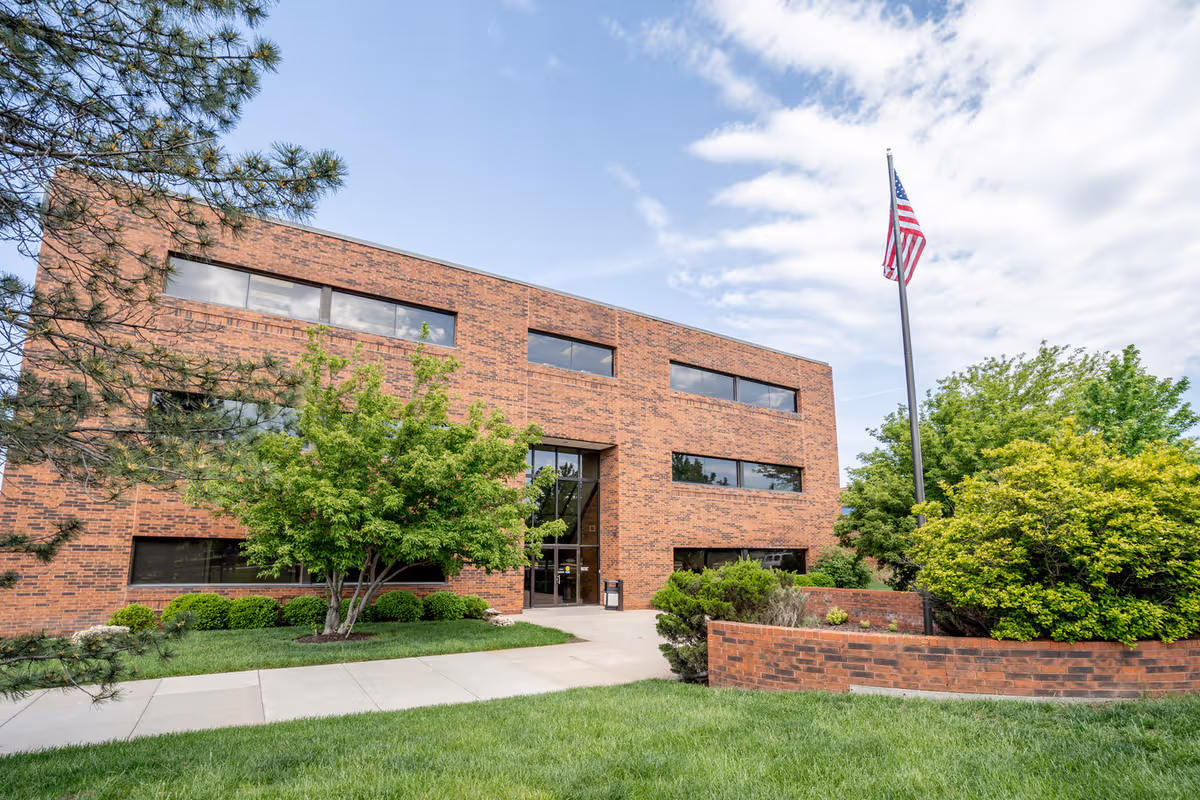 Brick two-story building with large windows, a central entrance, manicured lawn, and an American flag.