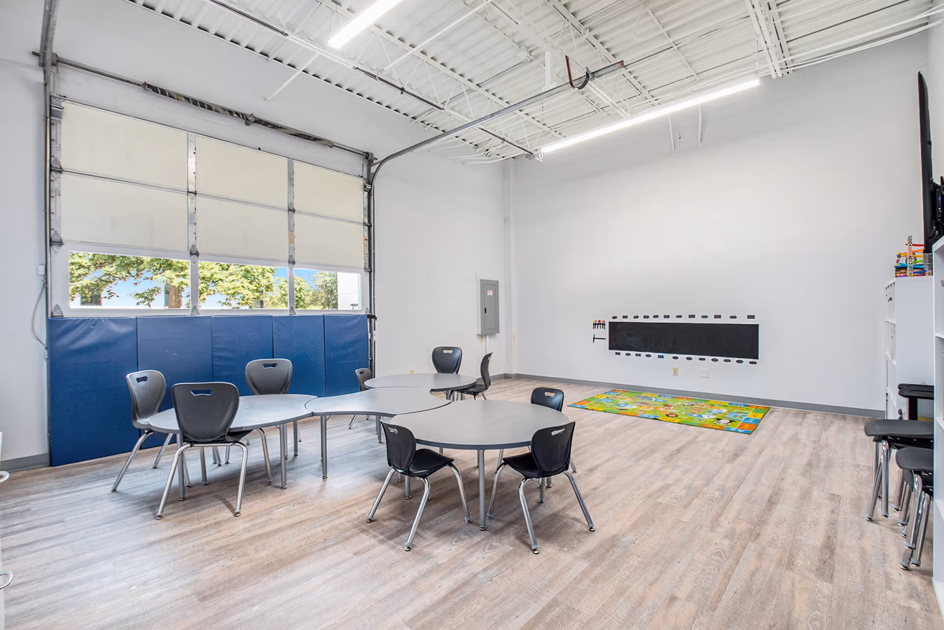 A spacious activity room with round tables, chairs, blue wall padding, and a colorful rug.