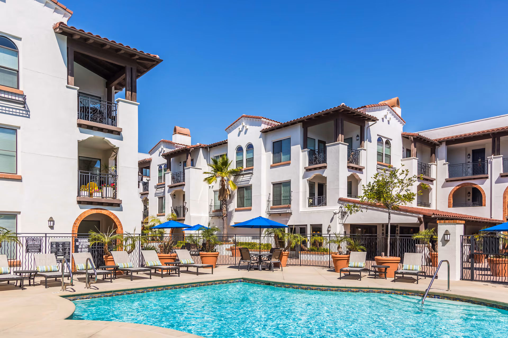 Outdoor swimming pool area with lounge chairs and blue umbrellas in front of a white multi-story building with balconies and terracotta roof tiles under a clear blue sky.