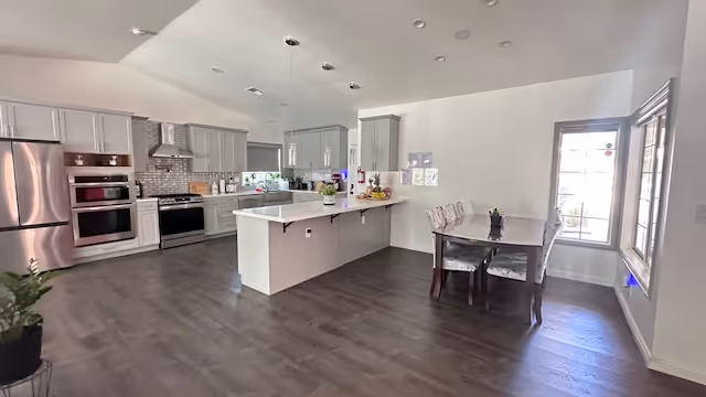Open kitchen with island, stainless steel appliances, and a small dining table by a window.