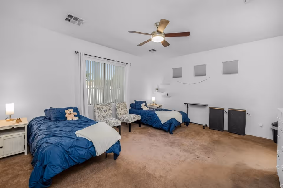 A bright bedroom with two twin beds, blue comforters, and a ceiling fan.