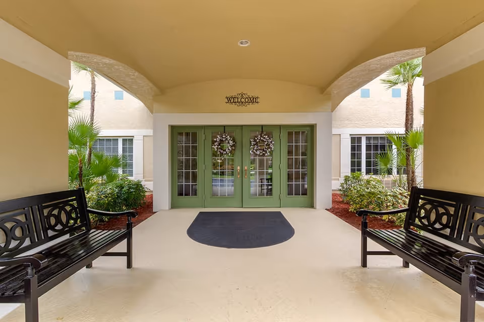 Covered entrance with green double doors decorated with wreaths, benches on either side, and a 'Welcome' sign above.