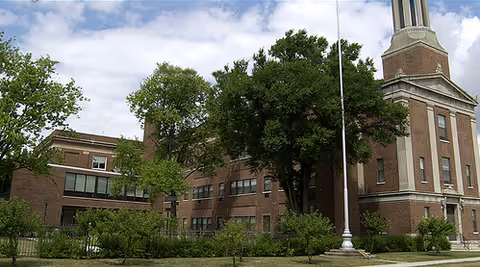 Brick senior living facility exterior with trees, a flagpole, and a tall turret.