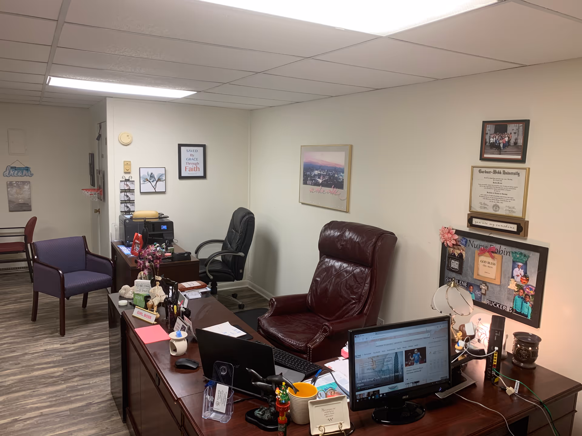 Office space with desk, laptop, chairs, and framed certificates on light walls.