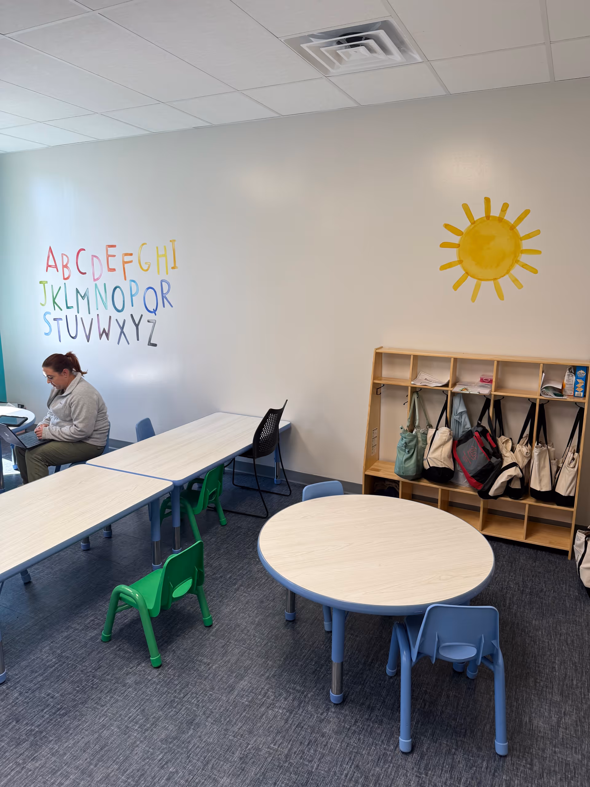 Interior classroom space featuring alphabet mural, sun illustration, cubbies, and small tables with chairs.