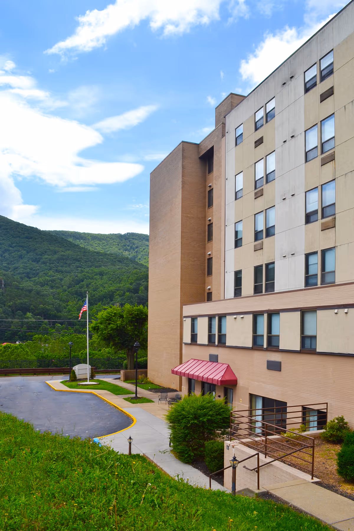 Exterior view of a multi-story building with beige and brown walls, featuring several windows and a red awning over the entrance. The building is surrounded by greenery, including bushes and a grassy hill, with a paved driveway and an American flag on a flagpole. In the background, there are forested hills under a partly cloudy blue sky.