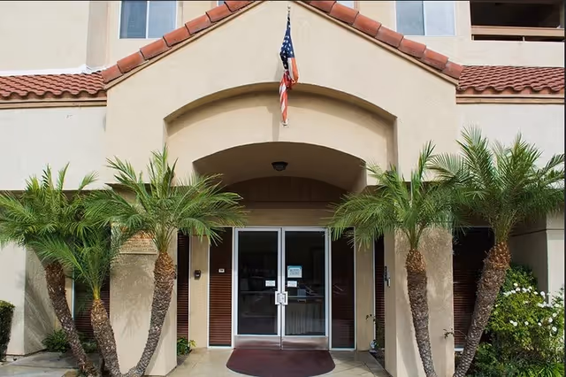 Entrance of a building with double glass doors under an archway, flanked by four palm trees and an American flag hanging above the entrance.