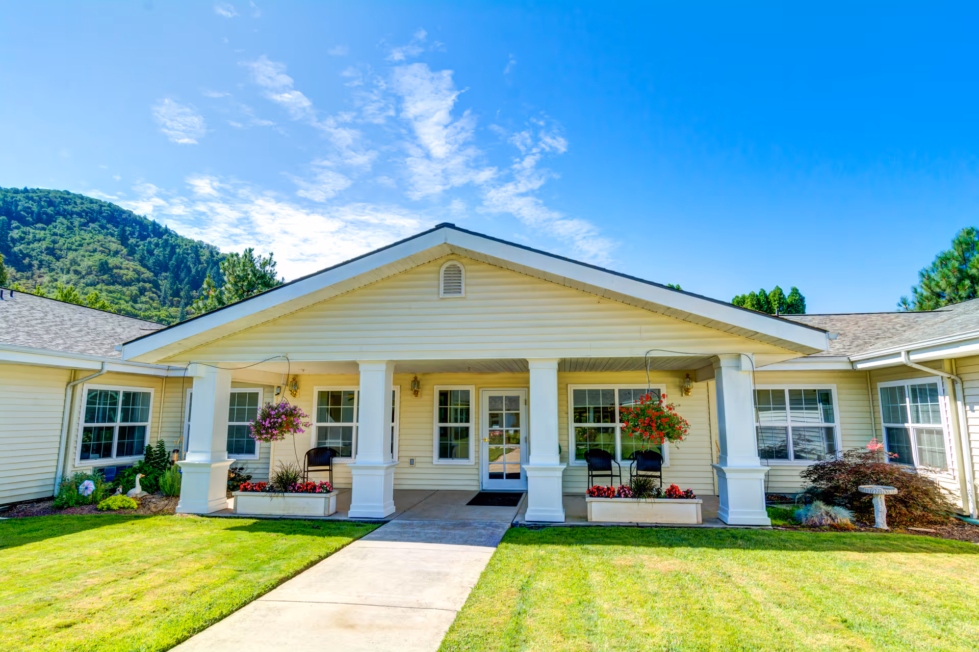 Front exterior view of Morrow Heights Assisted Living facility with a covered entrance supported by white columns, hanging flower baskets, and a well-maintained lawn under a bright blue sky with some clouds and a forested hill in the background.