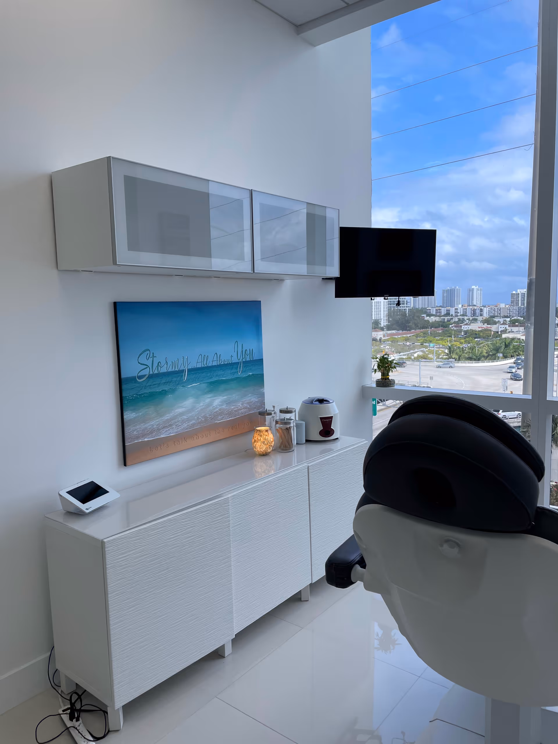 Bright, modern treatment room featuring a white cabinet, TV, and a reclining chair by large windows.