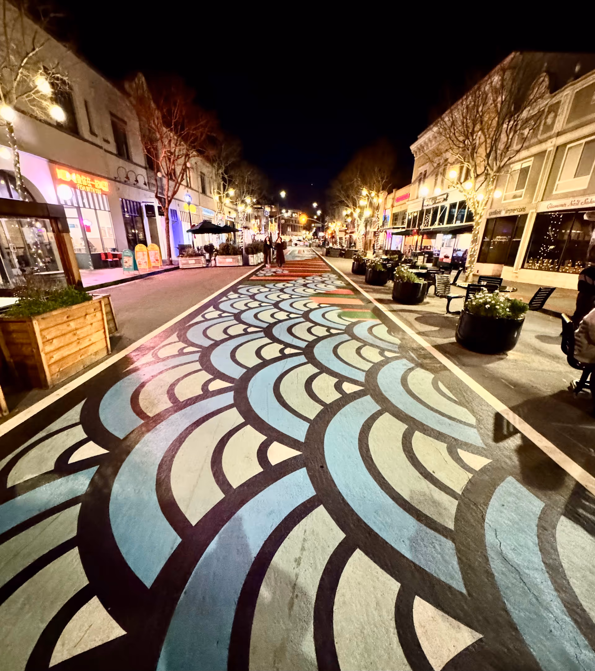 Nighttime street with colorful patterned pavement, storefronts, planters, and pedestrians downtown.