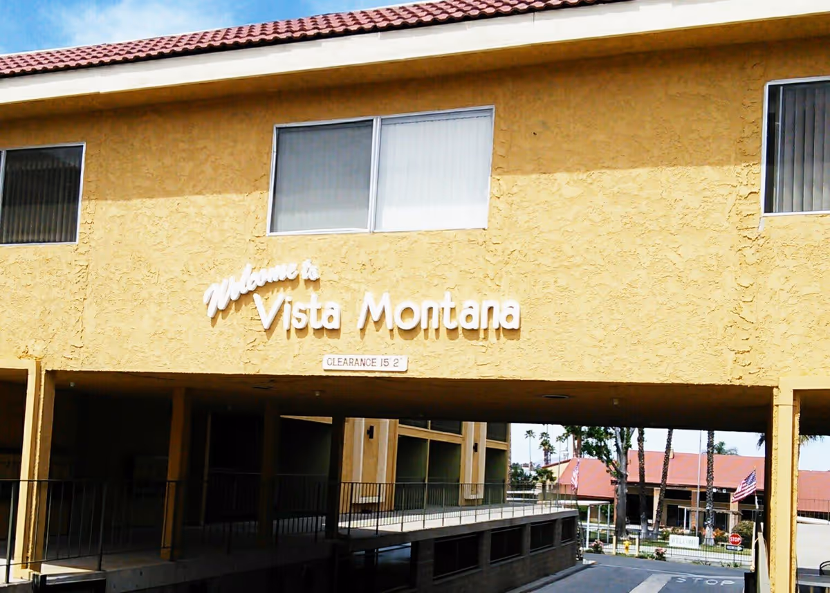 Exterior view of a yellow stucco building with a red tile roof. The building has a sign that reads 'Welcome to Vista Montana' above a covered driveway with clearance of 15 feet 2 inches. There are windows with vertical blinds and a glimpse of palm trees and another building with a red roof in the background.