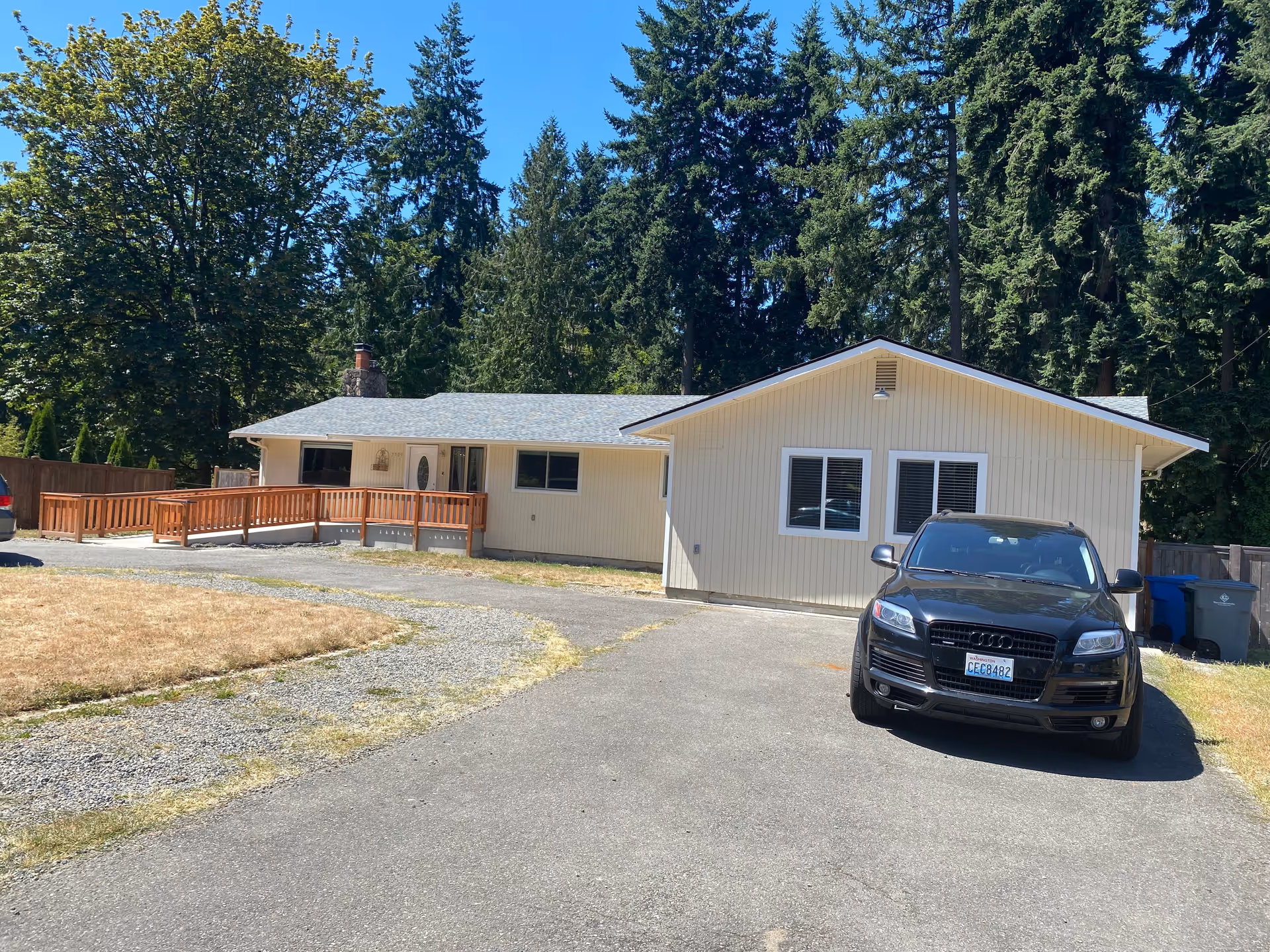 Exterior view of a single-story cream-colored building with a gray roof, surrounded by tall evergreen trees. There is a wooden ramp leading to the front door and a black Audi SUV parked on the driveway. The sky is clear and blue.