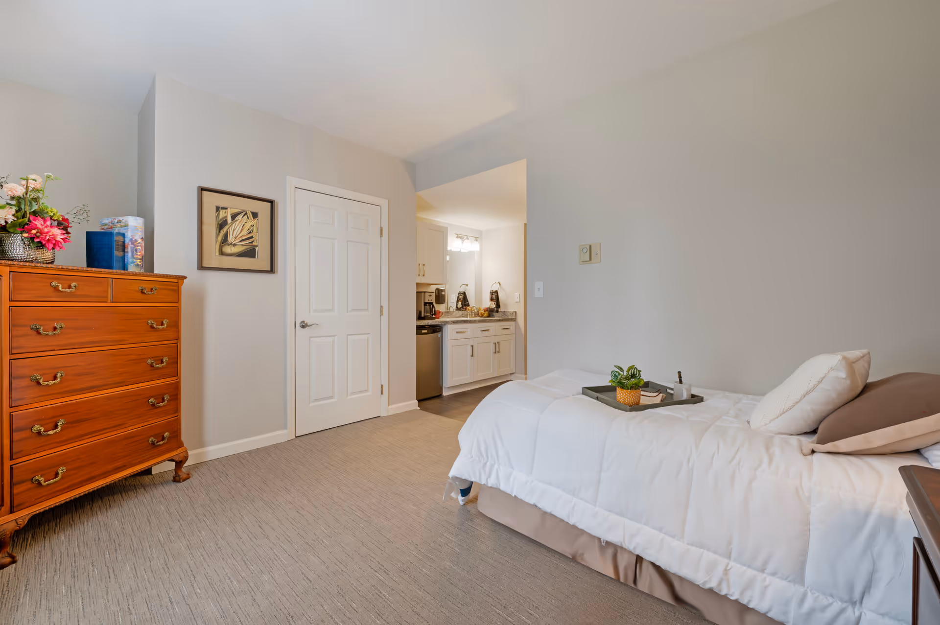 A tidy bedroom featuring a bed, wooden dresser, and doorway to a kitchenette.