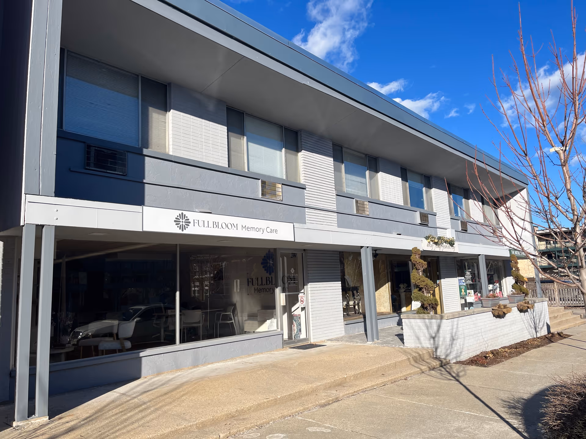 Two-story gray building with Full Bloom Memory Care sign and large storefront windows.