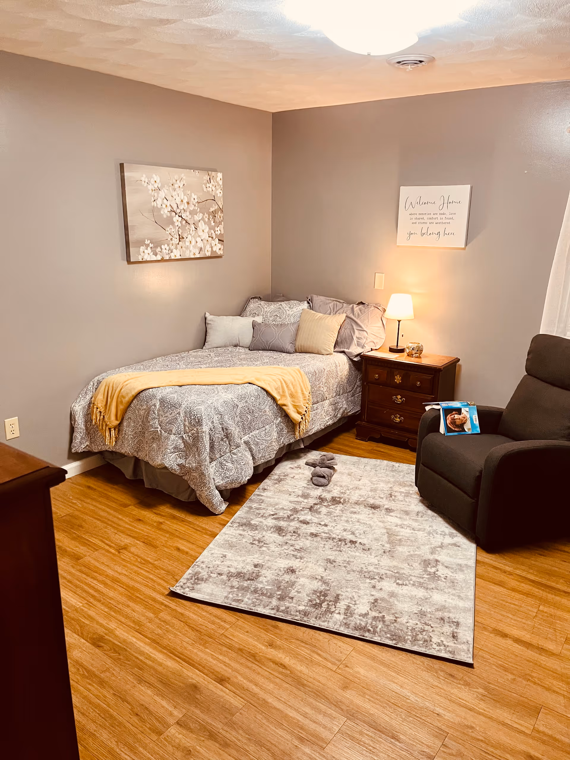 A cozy bedroom with a single bed covered in a patterned gray and white comforter, accented with multiple pillows and a yellow throw blanket. Next to the bed is a wooden nightstand with a small lamp and decorative items. A gray recliner chair is positioned near the nightstand, with a magazine resting on one arm. The floor is wooden with a light-colored area rug in front of the bed. The walls are painted a soft gray, decorated with a floral painting and a framed sign that reads 'Welcome Home'.