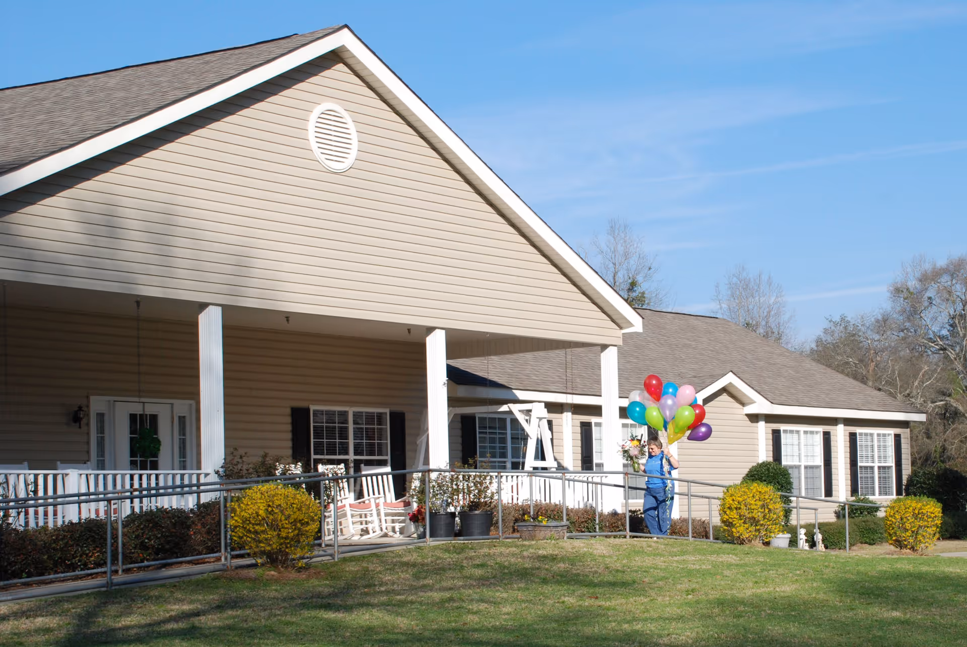 Exterior view of a single-story senior living facility building with beige siding and a covered porch. The porch has white railings, rocking chairs, and potted plants. A person holding colorful balloons and flowers is walking on the ramp leading to the porch. The sky is clear and blue, and there is green grass and bushes in the foreground.