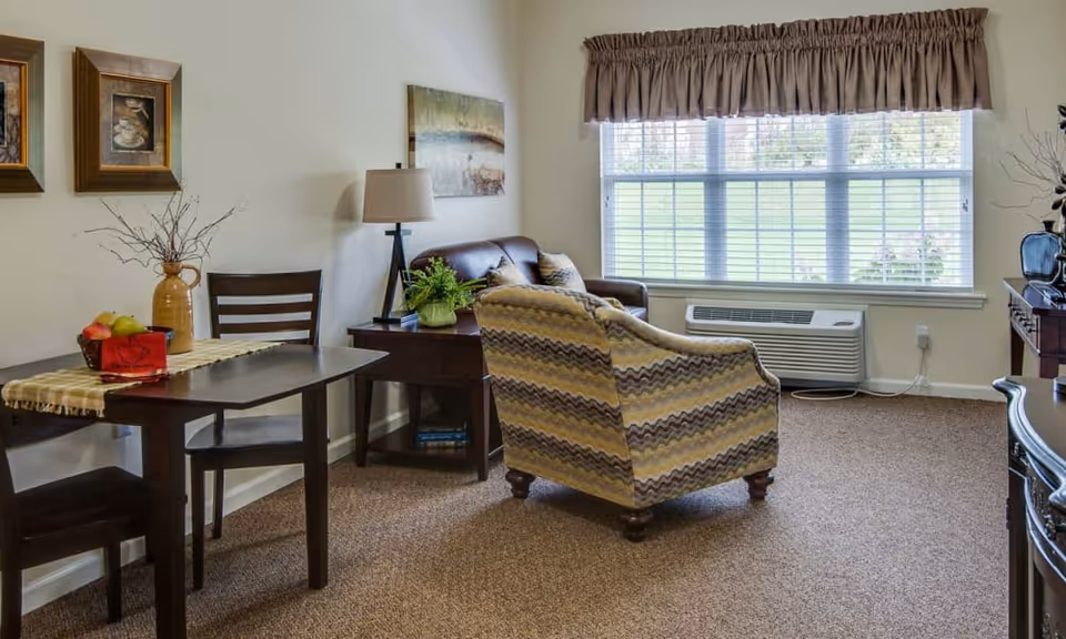 Sunlit living room with a patterned armchair, small dining table and chairs, side table with lamp, and a large window with an air conditioner.