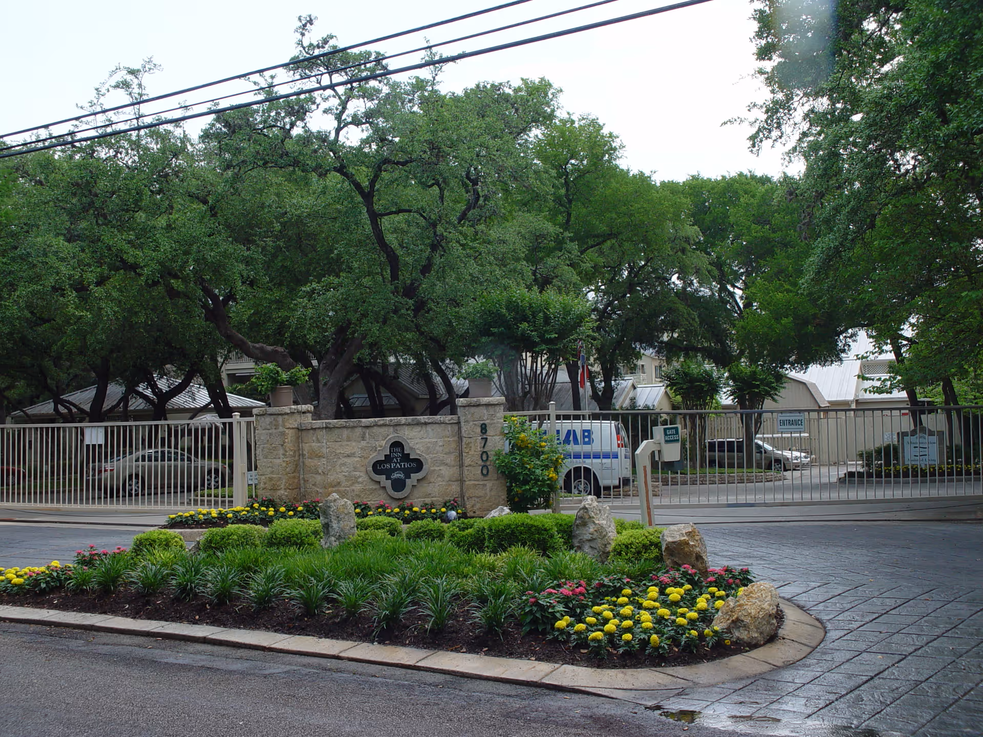 Gated entrance and landscaped circular driveway with a stone sign, metal fence, parked vehicles, and large trees.